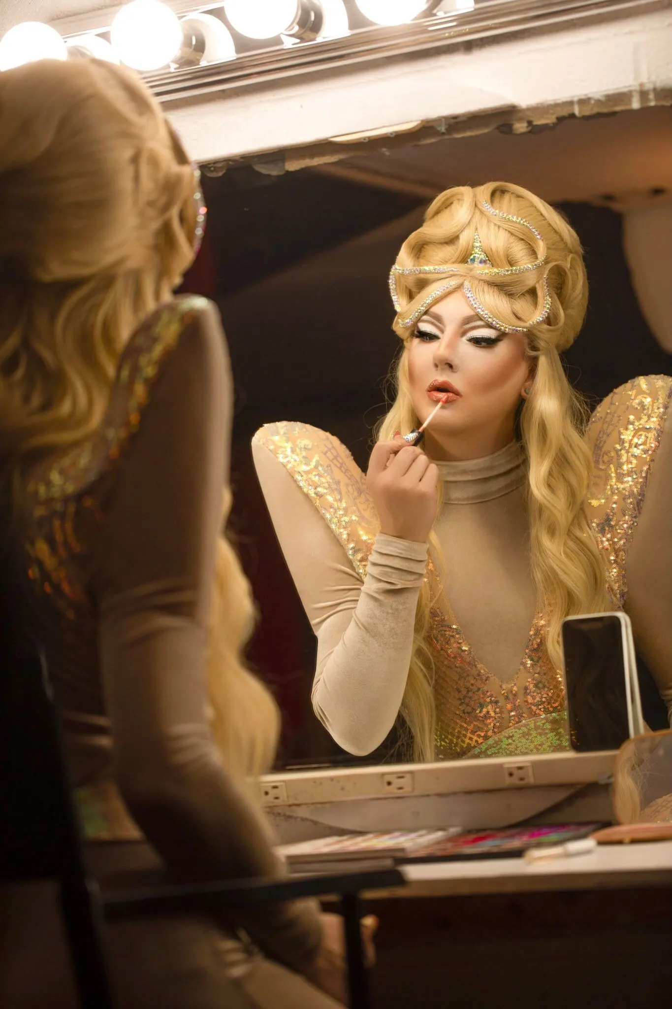 A queen in a gold crown headpiece and crystal bodice at a lit makeup mirror, lipstick in hand
