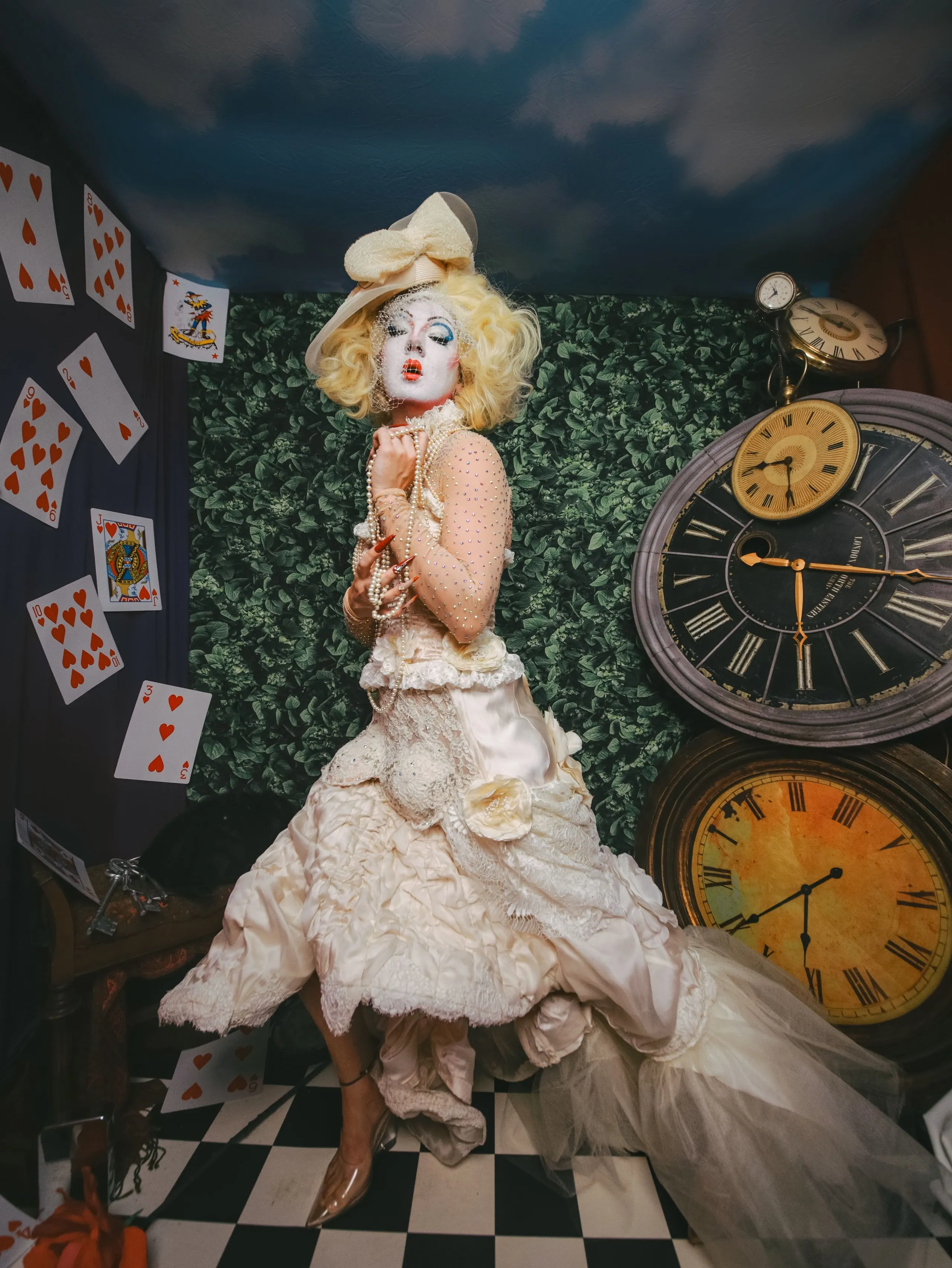 Figure in an ivory ruffled gown and bow hat on a checkered floor among giant clocks and playing cards