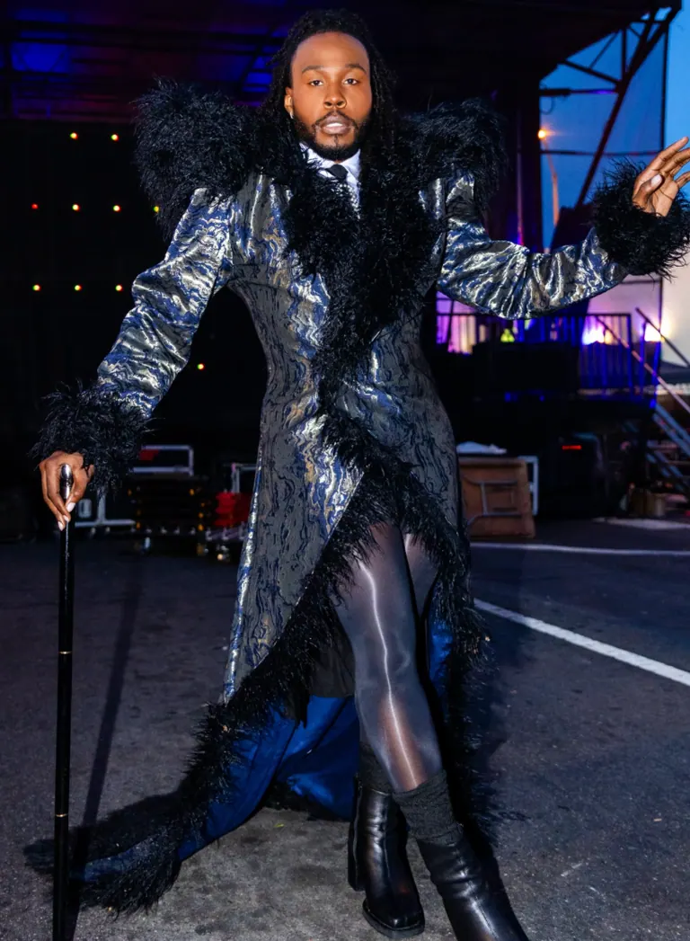 Performer in a silver snakeskin bodysuit reaching upward against a dark blue backdrop